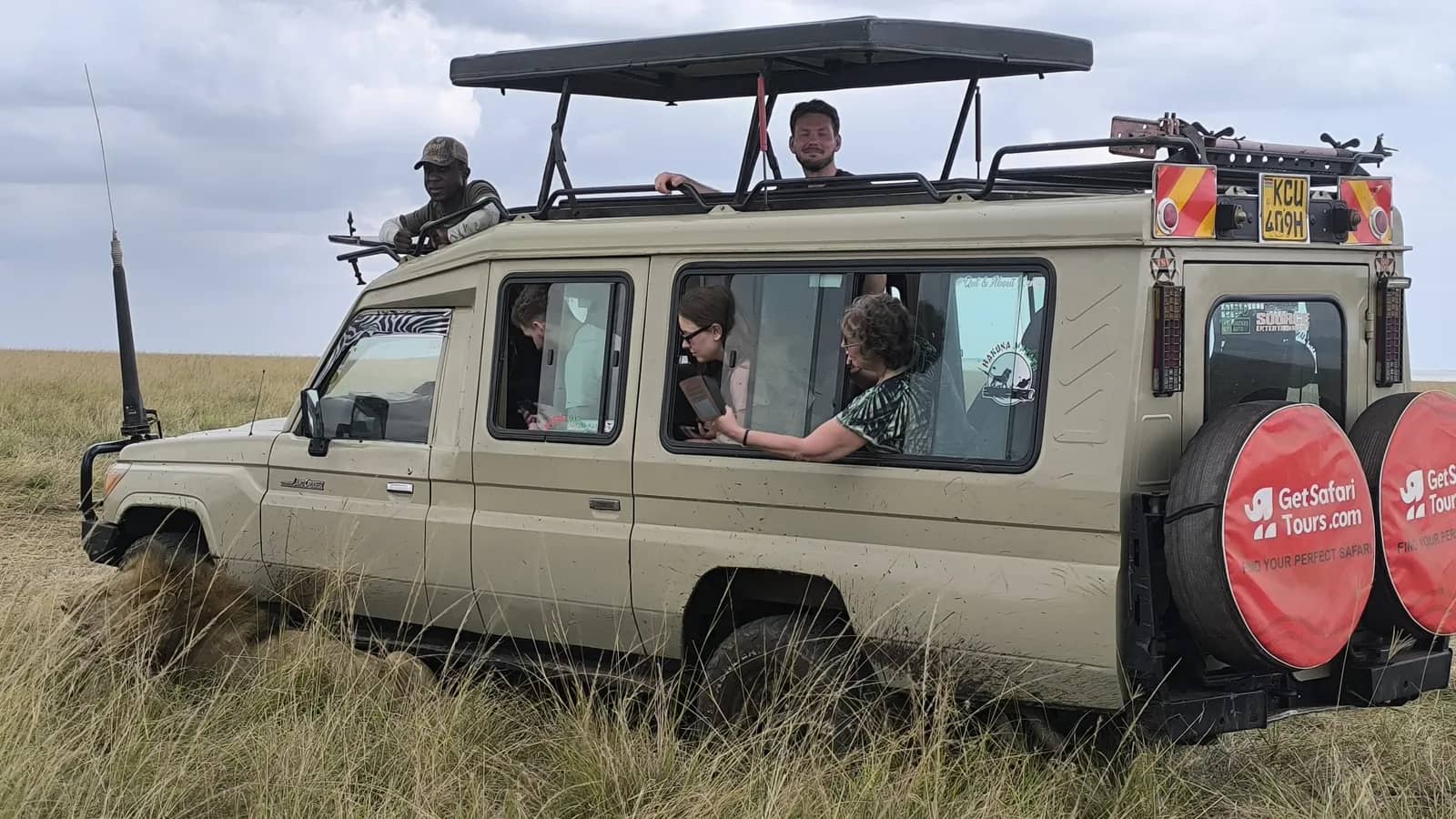 A lion resting in the grass next to a GetSafariTours safari vehicle with heads popped out of the roof